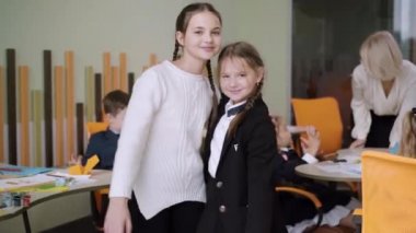 Güzel pozitif sınıf arkadaşlarının, kameraya sarılıp el sallamalarının portresi. Arka planda bulanık okul çocukları ve öğretmeniyle sınıfta poz veren ve gülümseyen iki beyaz kız öğrenci..