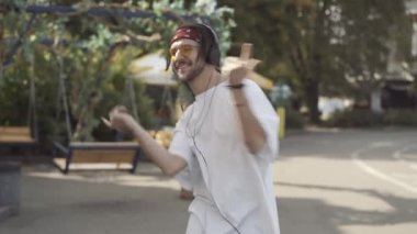 Rastalı Kafkas dövmeli genç adam güneşli şehir sokaklarında kulaklıkla müzik eşliğinde dans ediyor. Tasasız milenyum çocuğu portresi. Açık havada eğleniyor..