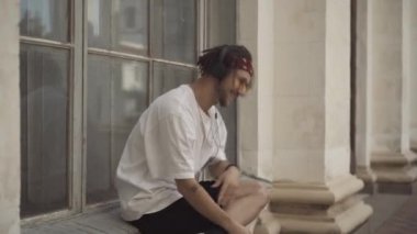 Rasta desenli sakallı, şehir penceresinde oturan ve kulaklıkla müziğin keyfini çıkaran adamın yan görüntüsü. Güneş gözlüklü kaygısız beyaz hippi dışarıda dinleniyor..