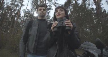 Dürbünle dışarıda duran iki mutlu ırklar arası motorcunun alt açısı. Gülümseyen Kafkasyalı kadın ve Orta Doğulu erkek bahar veya sonbahar ormanlarında çıkıyorlar. Sinema 4k ProRes Merkezi.