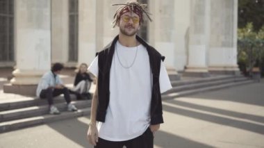 Güneş gözlüklü, gülümseyen hippi portresi. Arka planda oturan bulanık gotik arkadaşlarıyla kameraya bakıyor. Beyaz adam güneşli bir yaz gününde dışarıda poz veriyor.