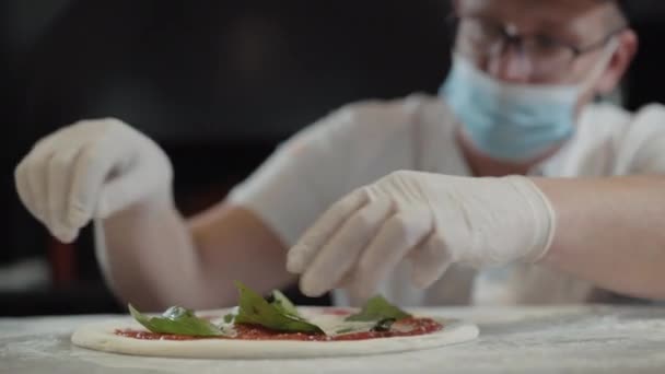 Zamazany kucharz w masce Covid-19 i rękawiczkach z dodatkiem liści oregano na pizzy. Profesjonalny kucharz kaukaski przygotowuje pyszne danie w kuchni restauracji na pandemię koronawirusa. — Wideo stockowe