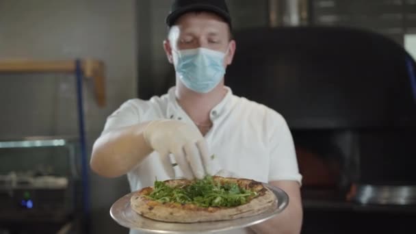 Chef professionnel caucasien en masque visage Covid-19 posant avec pizza prête dans la cuisine du restaurant. Homme flou tenant plat cuit savoureux ajoutant de la verdure et regardant la caméra. Affaires concernant le coronavirus. — Video