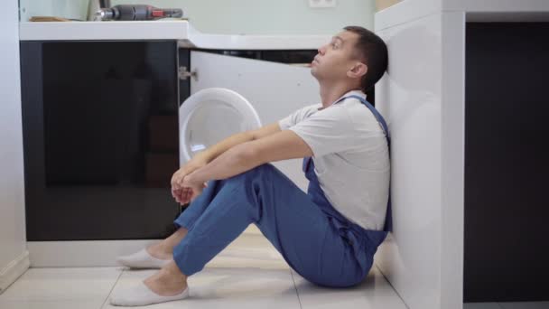 Portrait latéral de fatigué stressé jeune réparateur assis sur le plancher de la cuisine pensant. Frustré homme de service caucasien secouant la tête dans l'appartement avec des meubles cassés. Concept d'emploi. — Video