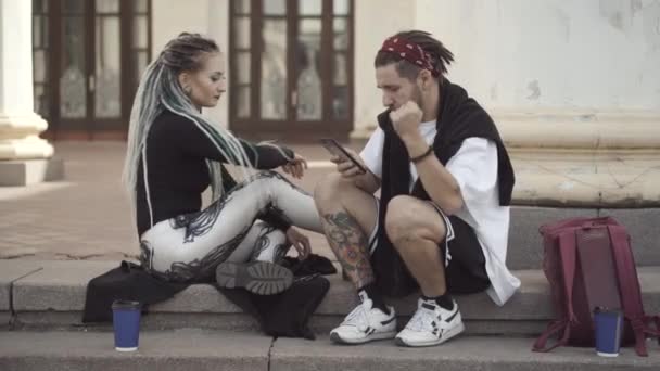 Quelques hippies détendus assis sur les escaliers urbains écoutant la musique du smartphone et parlant. Portrait de jeune homme et femme caucasien insouciant avec dreadlocks, tatouages et piercings à l'extérieur. — Video