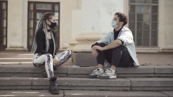 Un par de hippies modernos sentados en escaleras urbanas en Covid mascarillas tocando codos. Joven novio caucásico y novia citas en el día soleado en la ciudad en pandemia coronavirus. — Vídeo de stock