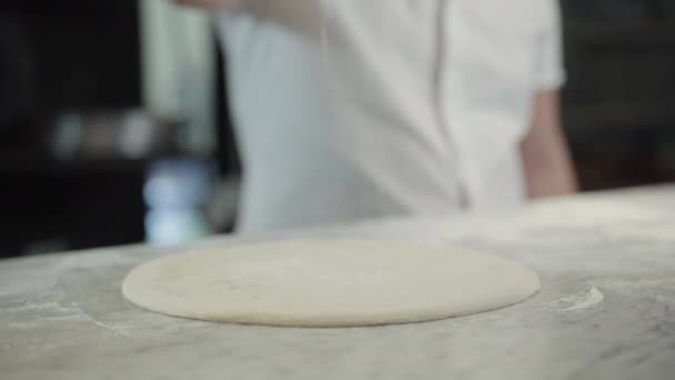 Chef caucásico irreconocible vertiendo harina sobre masa de pizza redonda en el restaurante. Hombre cocinero caucásico cocina sabroso plato nacional italiano en la cocina. Cocina y concepto culinario. — Vídeo de stock