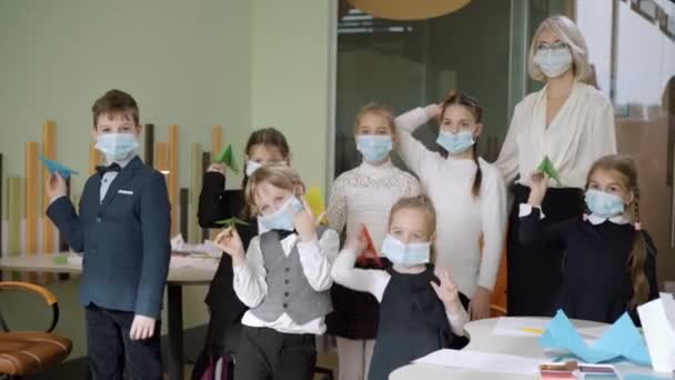 Lindos niños caucásicos en máscaras faciales Covid lanzando aviones de papel en el aula. Grupo de escolares divirtiéndose con el maestro en la escuela pública durante el bloqueo pandémico del coronavirus. — Vídeos de Stock