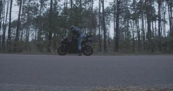 Extreme Weitwinkelaufnahme eines selbstbewussten Radfahrers, der die leere Straße entlangläuft, auf einem Motorrad sitzt und wegfährt. Junger Mann in Lederjacke und Motorradhelm. Cinema 4k ProRes Hauptquartier. — Stockvideo