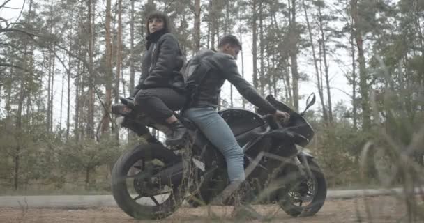 Jeune couple interracial de motocyclistes confiants assis dos à dos sur le vélo. Vue latérale large de l'homme du Moyen-Orient et de la femme blanche reposant sur le bord de la route. Siège social Cinema 4k ProRes. — Video