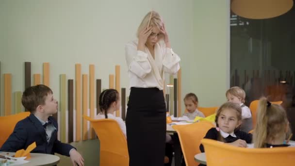 Épuisé jeune belle femme tenant la tête dans les mains comme des écoliers criant autour. Portrait d'une enseignante caucasienne fatiguée sur-stressée au travail à l'école primaire publique. Concept pédagogique. — Video