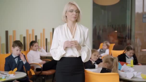 Preciosa maestra caucásica mirando alrededor del aula con los niños sentados en los escritorios, y sonriendo a la cámara. Retrato de una hermosa joven con anteojos que trabaja en la escuela pública. — Vídeos de Stock