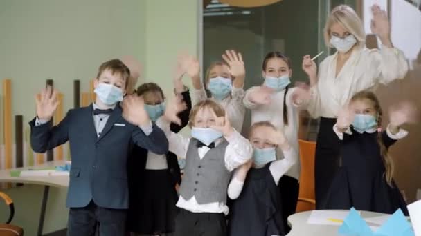 Grupo de escolares positivos y profesor en Covid máscaras faciales saludando a la cámara y sonriendo. Niños y niñas alegres y lindos posando en la escuela pública durante el brote pandémico de coronavirus. — Vídeos de Stock