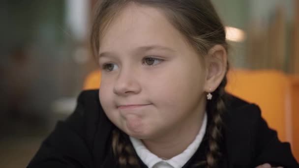 Close-up ansigt af lille keder skolepige tænker indendørs. Portræt af trist smuk kaukasisk brunette pige sidder ved skrivebordet i klasseværelset. Barndom og uddannelse koncept. – Stock-video