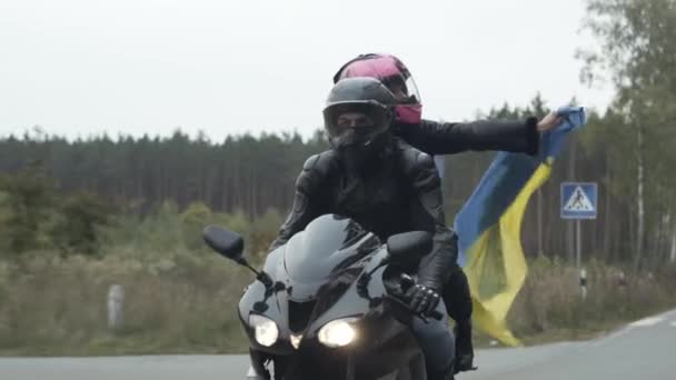 Confiado feliz pareja de jóvenes ciclistas con bandera ucraniana de conducción de motocicletas a lo largo de la carretera. Retrato de hombre y mujer en cascos montados en moto en Ucrania en día nublado nublado. — Vídeos de Stock