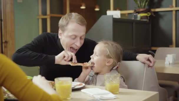 Fröhlicher kaukasischer Vater und seine Tochter beim Pizzaessen im Restaurant. Fröhlicher junger Mann und nettes Mädchen genießen das Abendessen in der Pizzeria, während die Frau vorne sitzt. Glückliche Familienfreizeit. — Stockvideo