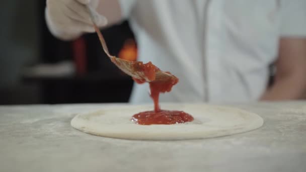 Male hand in glove spreading tomato sauce in pizza dough in restaurant kitchen. Unrecognizable Caucasian chef making delicious Italian dish indoors. Cook working in pizzeria. — Stock Video