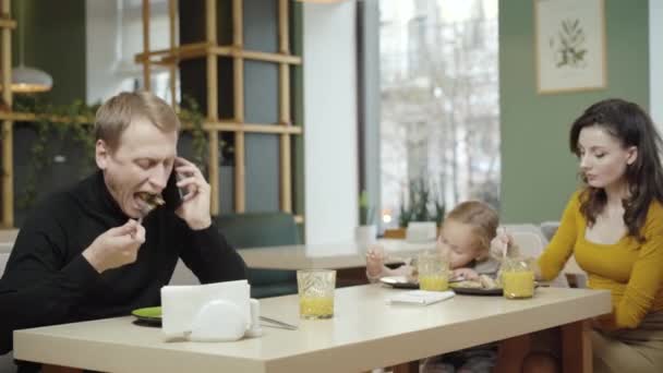 Zaneprázdněný běloch mluví po telefonu s rodinou v pizzerii. Portrét seriózního obchodníka, jak jí pizzu jako rozmazaná manželka a dcera, jak si užívají jídlo v pozadí. Koncept životního stylu. — Stock video