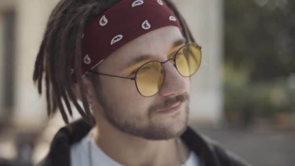 Cara de cerca del hombre milenario despreocupado con rastas que ponen gafas de sol y guiños. Retrato de positivo barbudo hippie caucásico coqueteando en la cámara en la soleada ciudad al aire libre. — Vídeo de stock