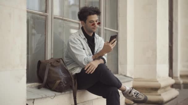 Впевнений, гарний брунатний хіпі-серфінг в інтернеті на смартфоні сидить на підвіконні. Портрет розслабленого кавказького чоловіка, який розмовляє на вулиці в місті. Сучасний спосіб життя. — стокове відео