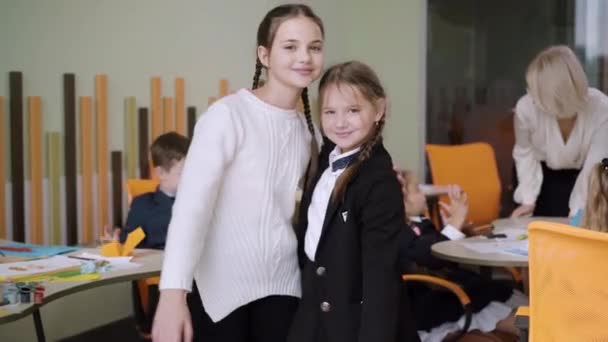 Porträt schöner positiver Klassenkameraden, die sich umarmen und in die Kamera winken. Zwei süße kaukasische Schulmädchen posieren im Klassenzimmer mit verschwommenen Schulkindern und Lehrern im Hintergrund und lächeln. — Stockvideo
