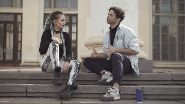 Aufgeregte junge kaukasische Hippie sitzt auf städtischen Treppen mit Gothic-Frau und redet. Porträt eines entspannten Millennial-Paares, das sich an einem sonnigen Sommertag im Freien trifft. Beziehung und Subkultur-Konzept. — Stockvideo