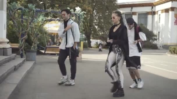 Vista lateral de tres jóvenes despreocupados bailando como caminando en la soleada ciudad de arriba. Columna blanca pasando por delante. Relajado caucásico hippie hombres y mujeres disfrutando de ocio al aire libre. — Vídeos de Stock