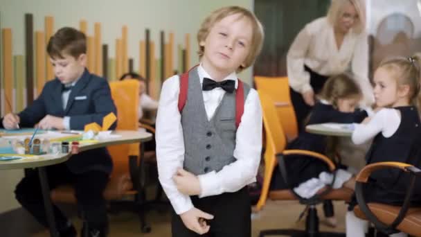 Buon scolaro che regola lo zaino e sorride alla telecamera in piedi in classe con insegnante offuscata e compagni di classe sullo sfondo. Ritratto di ragazzo caucasico carino positivo in posa nella scuola pubblica. — Video Stock