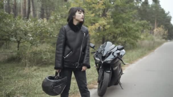 Stresowana młoda kobieta autostopem na pustej drodze z zepsutym motocyklem stojącym w tle. Smutny biały motocyklista ma problemy z motocyklem na zewnątrz. Koncepcja wypadków. — Wideo stockowe