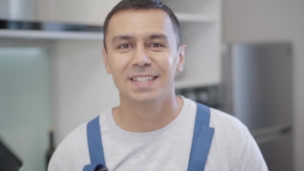 Cabeza del joven hombre de servicio morena con ojos marrones sosteniendo taladro eléctrico sonriendo mirando a la cámara. Primer plano retrato de maestro caucásico profesional posando en el interior con herramienta de reparación. — Vídeos de Stock