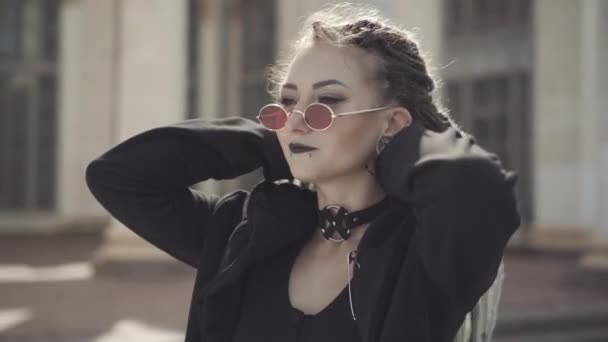 Primer plano de mujer hippie confiada con piercings y rastas en gafas de sol mirando a la cámara sonriendo. Retrato de una joven dama caucásica posando al aire libre en la ciudad urbana. Estilo de vida y subcultura. — Vídeo de stock