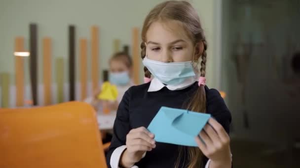 Retrato de colegial caucasiana triste em Covid-19 máscara facial lançando avião de papel em sala de aula. Pequena menina bonita caucasiana ter pausa durante o estudo sobre pandemia de coronavírus. — Vídeo de Stock