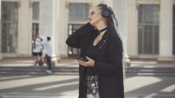 Mulher hippie caucasiana despreocupada em óculos de sol e fones de ouvido dançando na ensolarada rua da cidade com homens embaçados falando ao fundo. Retrato de jovem gótico relaxado desfrutando de música ao ar livre. — Vídeo de Stock