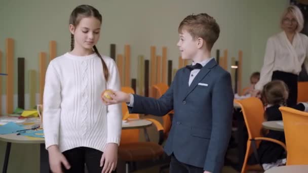 Lachende schooljongen deelt appel met schoolmeisje in de klas. Liefdevolle kleine blanke jongen die fruit geeft aan een mooi meisje op de openbare school. Romantiek en kindergeflirt. Eerste liefde concept. — Stockvideo