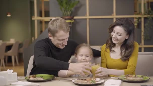 Joyeux jeune famille cliquetis avec du jus d'orange dans le restaurant de pizza et souriant à la caméra. Portrait de joyeuse mère caucasienne, père, et mignonne petite fille reposant dans la pizzeria. — Video