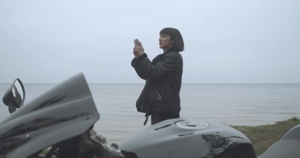 Triste donna caucasica che cerca la rete mobile sulla riva del fiume. Ritratto di giovane bella motociclista che viaggia in moto nella nuvolosa giornata primaverile o autunnale. Cinema 4k ProRes HQ. — Video Stock