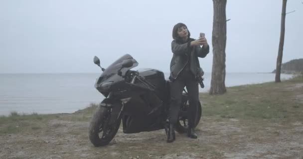 Ampio ritratto di una bella motociclista donna che fa selfie all'aperto sulla riva del fiume. Giovane donna caucasica affascinante e sicura di sé che fotografa sullo sfondo del paesaggio. Cinema 4k ProRes — Video Stock