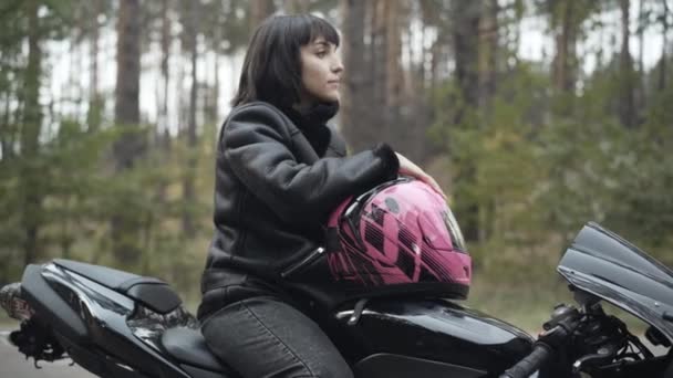 Vista laterale ritratto di fiduciosa donna caucasica sorridente seduta su moto nera con casco rosa e distogliendo lo sguardo. Bella giovane motociclista donna pensando all'aperto. Stile di vita e hobby. — Video Stock