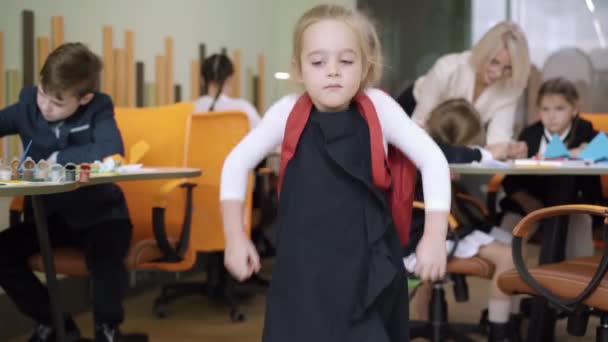 Niña extremadamente hermosa de pie con mochila en el aula con compañeros de clase borrosos y maestro en el fondo. Retrato de linda positiva colegiala caucásica posando y sonriendo a la cámara. — Vídeo de stock