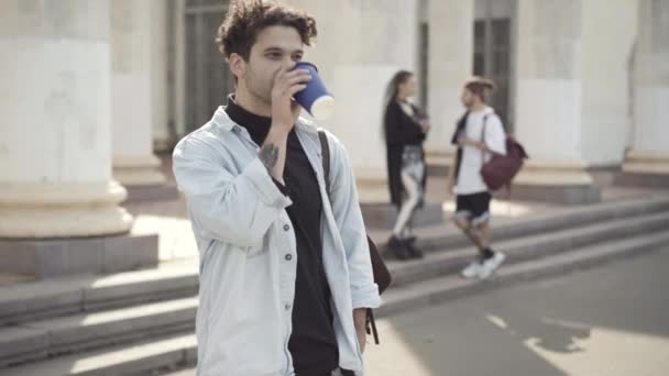 Tattooed curly-haired Caucasian man drinking coffee and smiling at camera with blurred hippie friends talking at background. Portrait of positive young guy posing outdoors. Subculture concept. — Stock Video