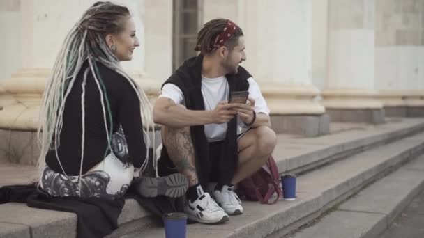 Porträt eines fröhlichen Hippie-Freundes, der auf städtischen Treppen sitzt und mit Freundin spricht. Entspanntes junges kaukasisches Millennial-Paar genießt den sonnigen Tag im Freien. Modernes Beziehungskonzept. — Stockvideo
