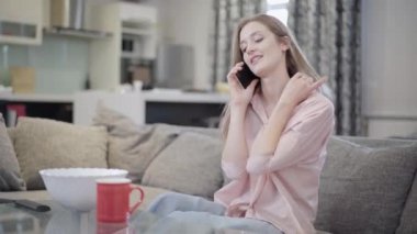 Oturma odasındaki koltukta oturan kendine güvenen pozitif kadın telefonda konuşuyor. Hafta sonları evde dinlenen güzel beyaz bir bayanın portresi. Mutluluk ve yaşam tarzı kavramı.