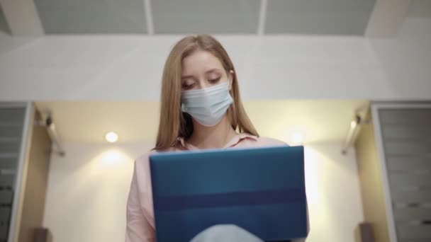 Egy beteg fiatal nő arcképe Covid-19-es maszkban, laptoppal és köhögéssel. Gyönyörű, karcsú hölgy szörfözik az interneten, elszigetelten otthon, a hálószobában. Coronavirus pandémiás fogalom. — Stock videók