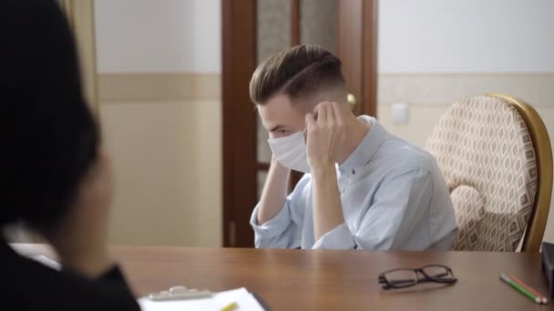 Vue de côté portrait d'un patient triste homme portant un masque facial Covid assis dans un bureau de psychologue. Portrait d'un homme caucasien désespéré en consultation psychologique sur le confinement en cas de pandémie de coronavirus. — Video