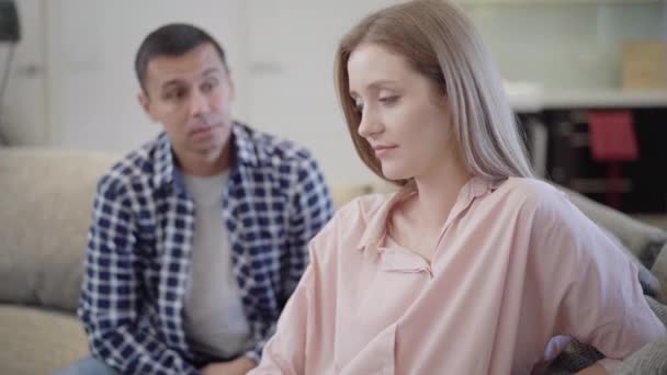 Triste jeune belle femme assise sur le canapé comme un homme caucasien flou parlant à l'arrière-plan. Portrait de petite amie malheureuse écoutant petit ami désolé pour l'infraction. Difficultés relationnelles. — Video