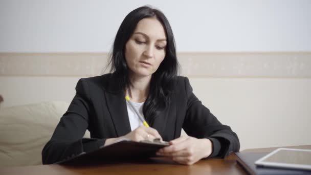 Retrato de mujer caucásica confiada hablando y escribiendo síntomas en papel. Psicóloga mujer concentrada consultando en interiores. Profesión psicológica y concepto de estilo de vida. — Vídeo de stock