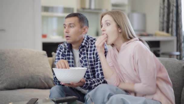 Sidovy av absorberade unga par tittar på skräckfilm och äter popcorn hemma. Porträtt av tusenåriga kaukasiska man och kvinna tittar på TV på helgerna i vardagsrummet. — Stockvideo