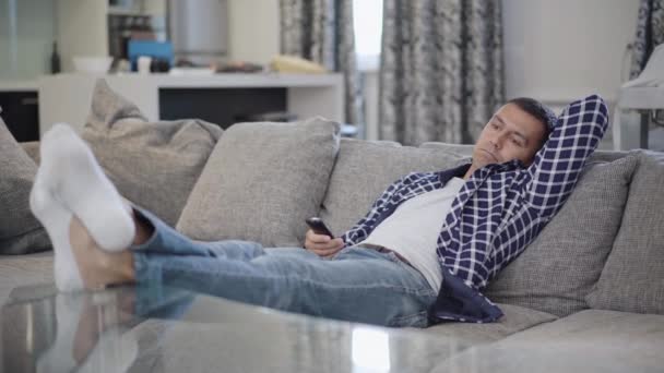 Ampla foto de homem caucasiano relaxado deitado em canais de troca de sofá com controle remoto. Retrato de um jovem bonito entediado a ver televisão em casa dentro de casa. Estilo de vida e lazer. — Vídeo de Stock