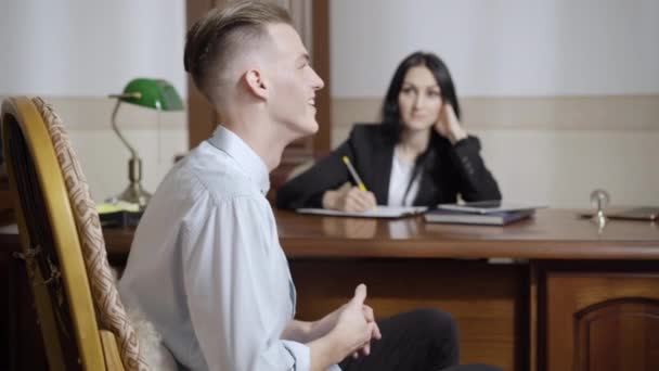 Side view of sick Caucasian young man talking and laughing out loud in psychologist office. Blurred concentrated woman listening patient at background. Mental illness and health concept. — Stock Video