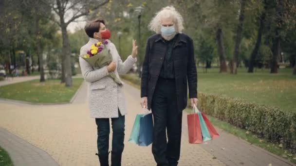 Široký záběr seniorského páru v Covid-19 obličejových maskách kráčejících s kyticí květin a nákupních tašek v jarním nebo podzimním parku a mluvících. Šťastný kavkazský muž a žena randění venku. — Stock video